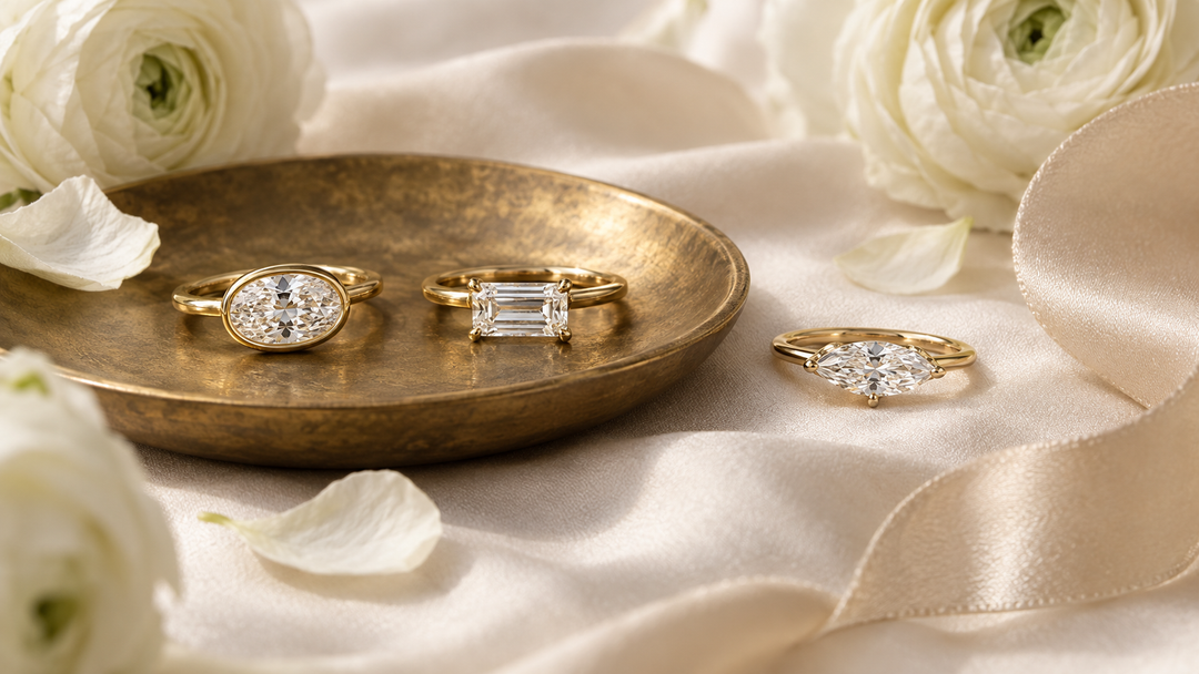 Three east-west engagement rings in yellow gold, an oval bezel solitaire, a sideways emerald cut, and a horizontal marquise, on ivory silk with a brass dish and white ranunculus